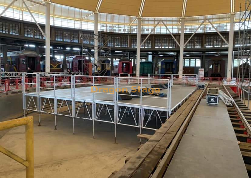  Pódio de trilhos de alumínio 12x8m com 2 escadas de acesso e rampa de carga/descarga ao longo do palco traseiro